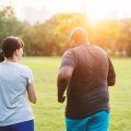 Two people, a man and woman, wear exercise gear outdoors in a green park and walk away from the camera.